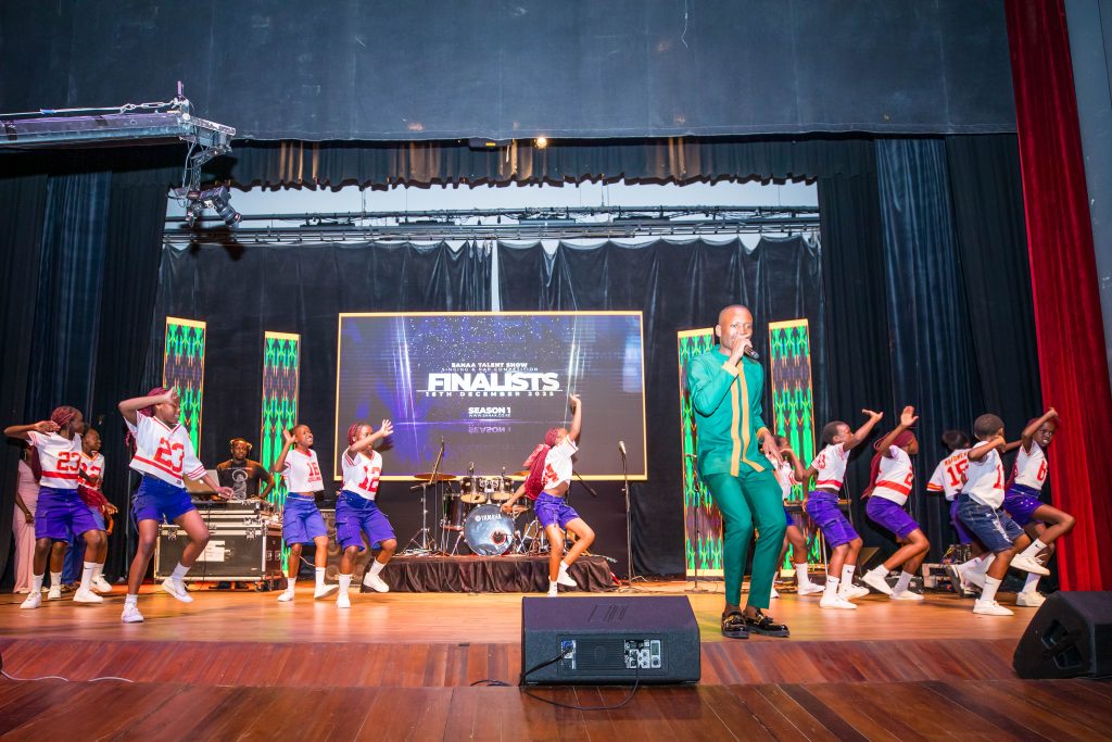 A group of young dancers in matching sports jerseys performing a choreographed routine alongside a lead singer in a green suit.
