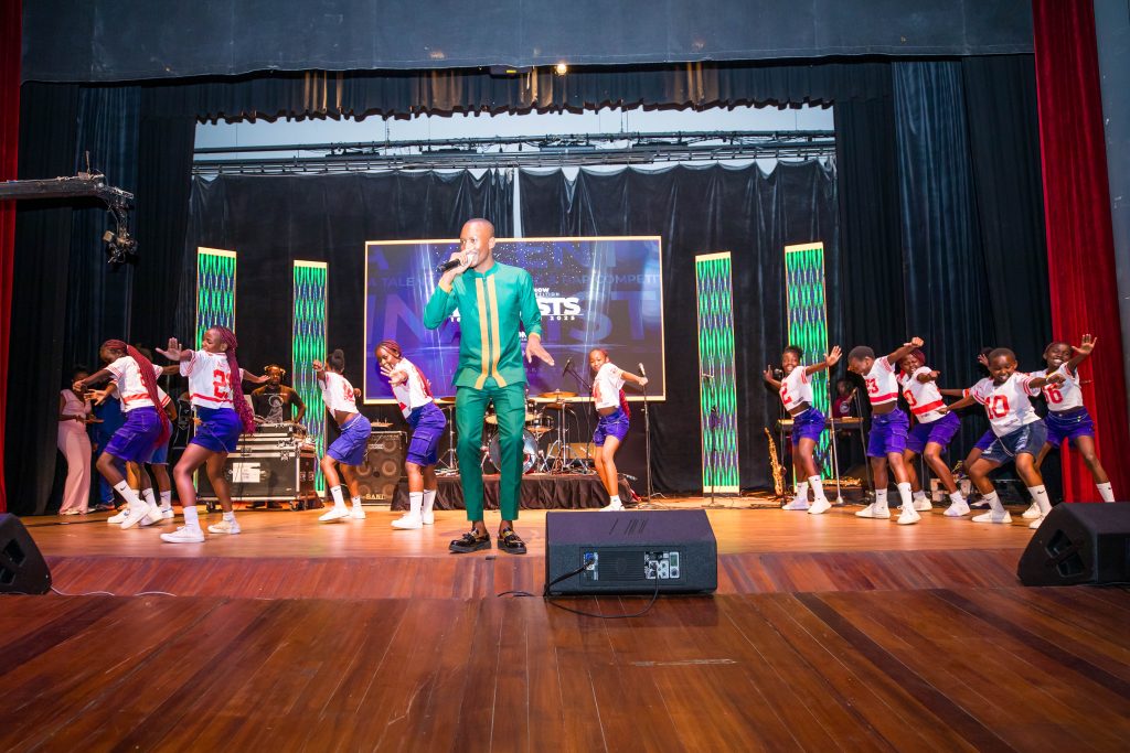 A large group of young dancers in white and red jerseys performing synchronized moves alongside a lead singer in a green suit.