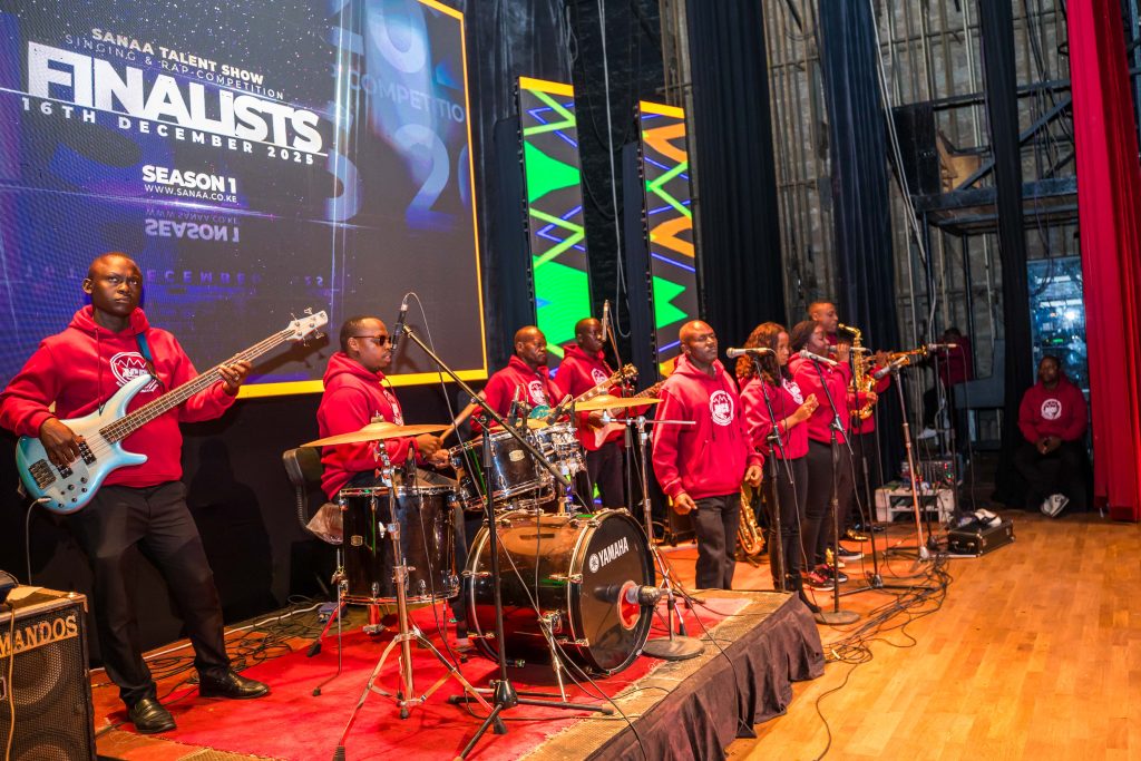 A full live band wearing matching red Mintglint Media hoodies performing on a stage with a digital screen in the background reading "Sanaa Talent Show Finalists."