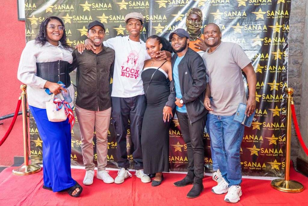 A diverse group of participants and guests smiling and posing in front of a black backdrop with the gold Sanaa Talent Show logo.