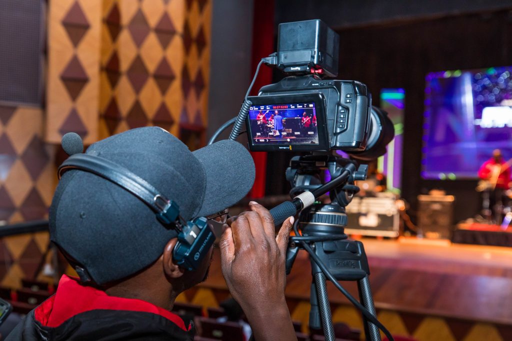A close-up of a professional video camera operator filming the stage; the camera screen shows a live feed of the performers.