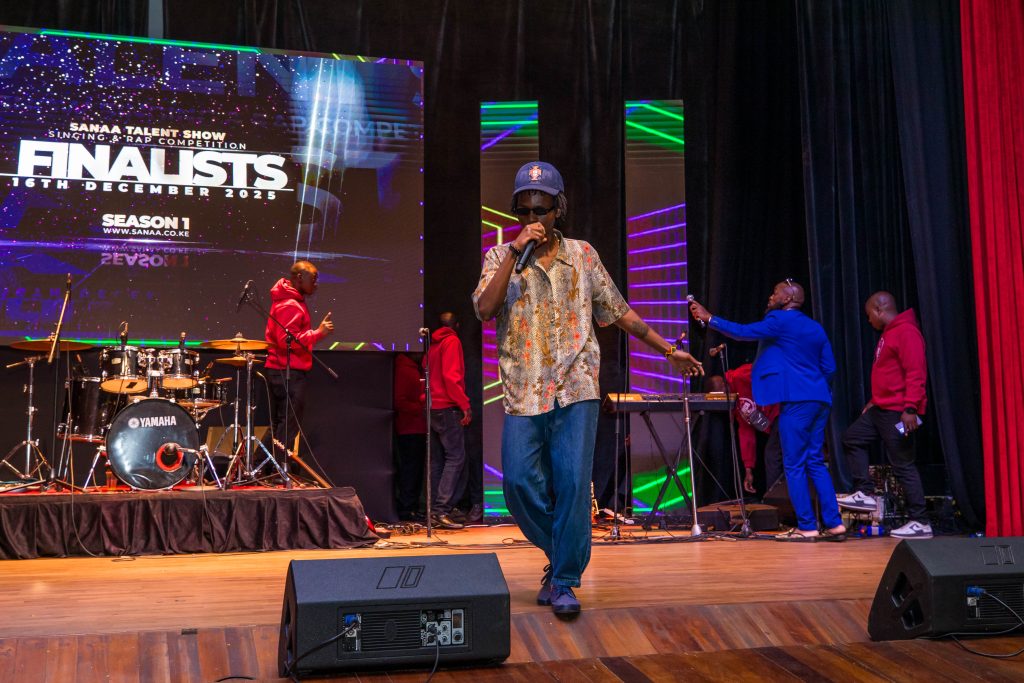 A male rapper in a patterned shirt, blue jeans, and a cap performing on stage with a microphone.