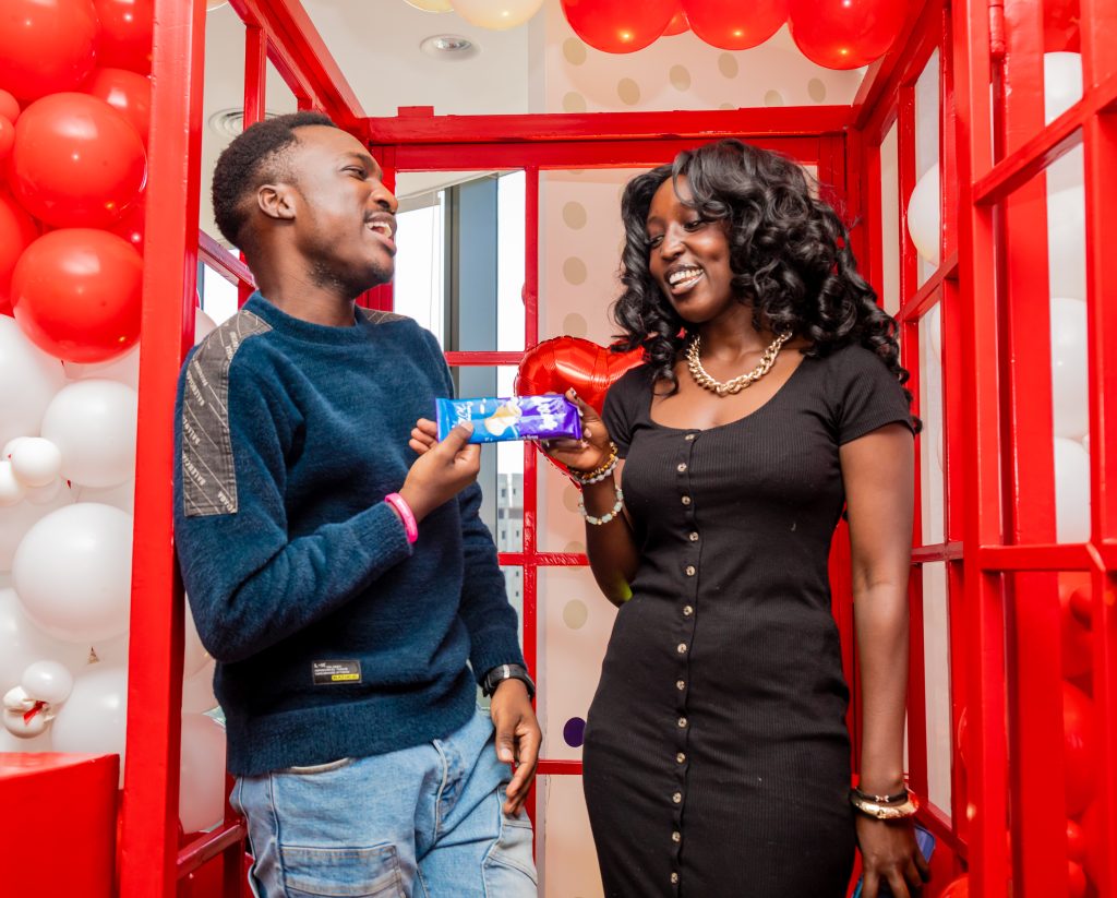 A man offering a chocolate bar to a laughing woman inside a red telephone booth prop during a Valentine's event.