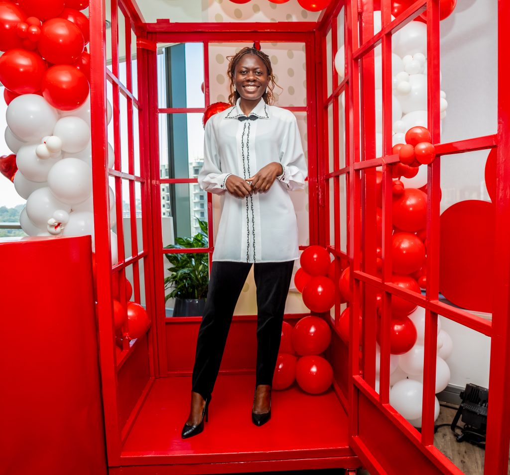 A woman in a white blouse posing elegantly inside a red telephone booth decoration surrounded by white and red balloons.