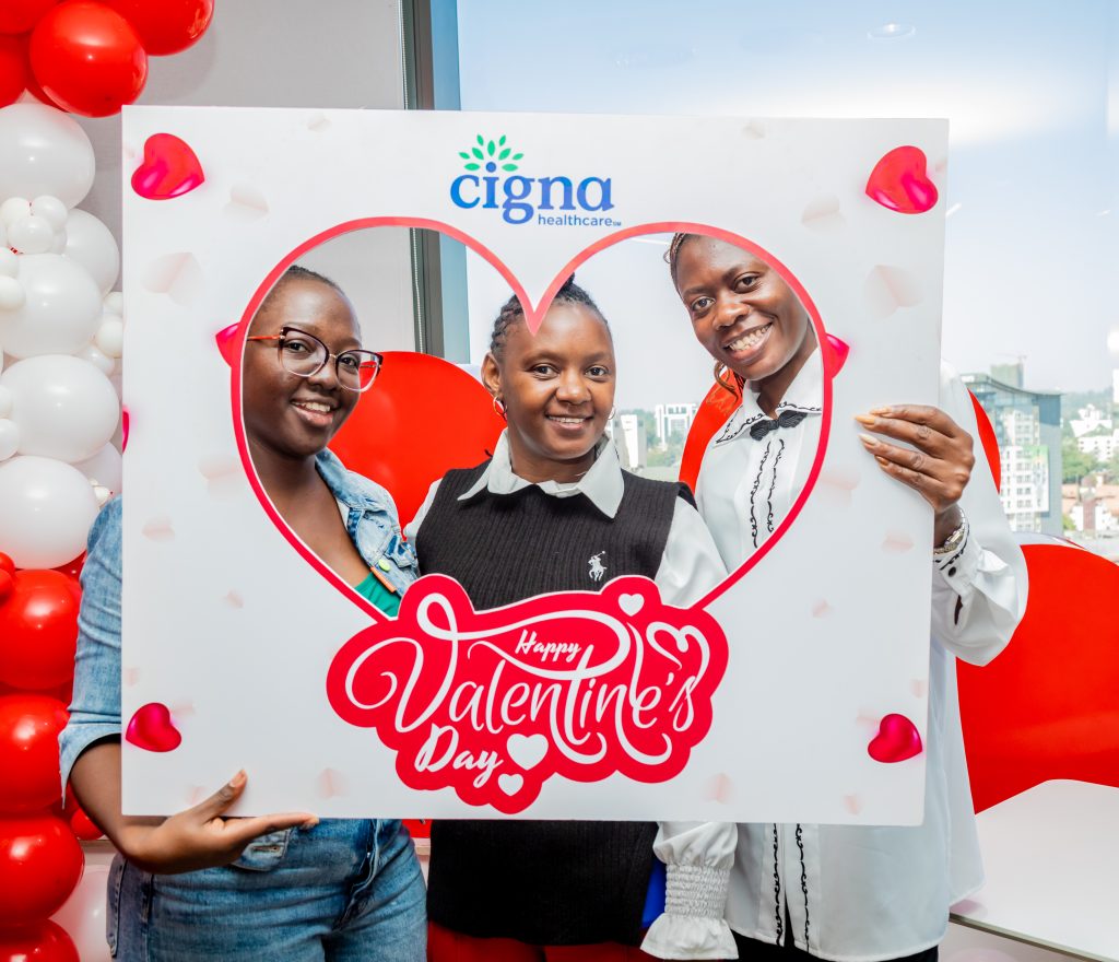 Three women smiling through a heart-shaped photo booth frame at a daytime office Valentine's celebration.