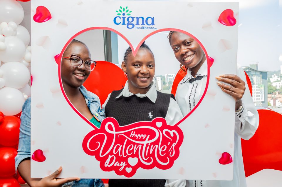 Three women smiling through a heart-shaped photo booth frame at a daytime office Valentine's celebration.