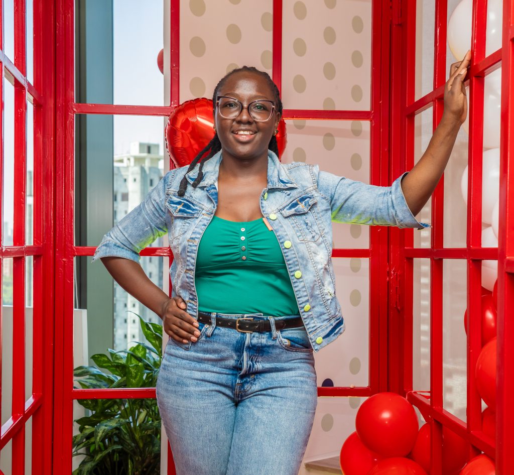 A woman in green posing playfully with hands raised in front of a large red "LOVE" sign and heart balloons.