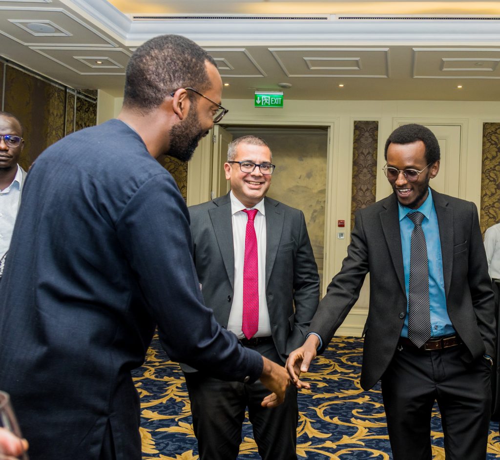 Two men shaking hands enthusiastically while another looks on smiling at a JBQ Africa event, covered by Mintglint Media.