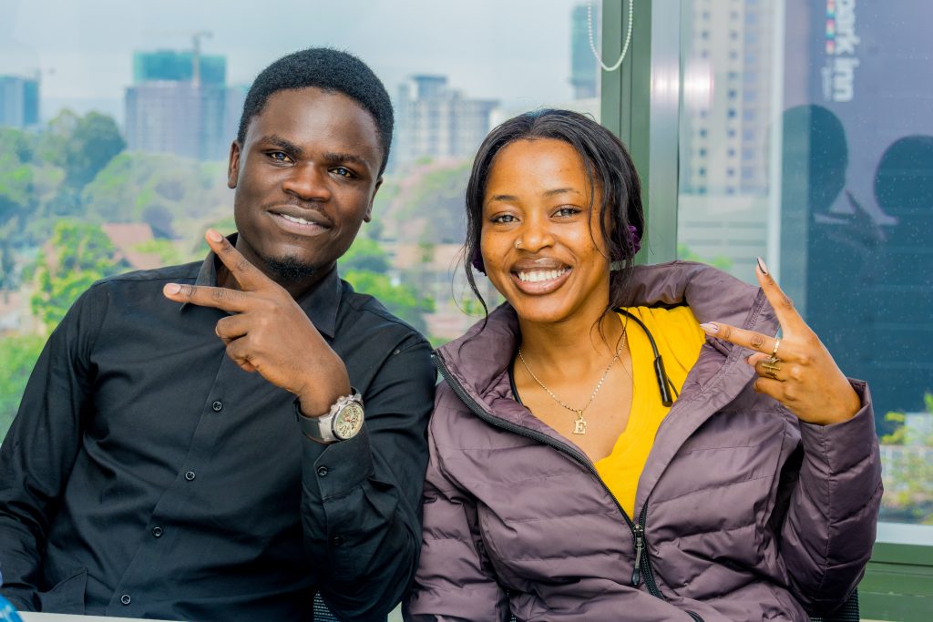 A male and female colleague smiling and throwing peace signs in front of a bright office window with the Nairobi skyline.