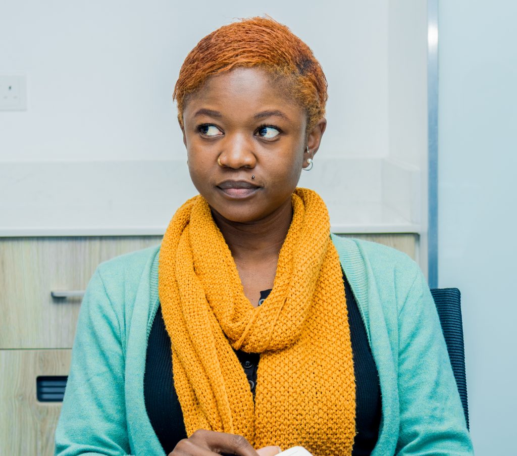 A woman with short orange hair and a chunky yellow scarf looking thoughtfully to the side during an office meeting.
