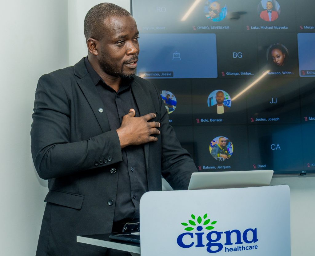 A man presenting at a Cigna podium with his hand on his chest, while a screen displays virtual attendees behind him.