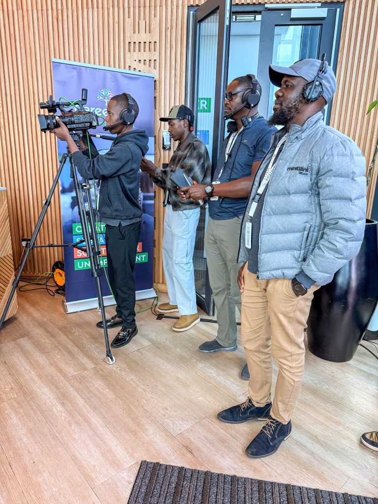 Three Mint Glint Media production crew members standing next to a Sony camera on a tripod. One operates the camera while the other two, wearing headsets and Mint Glint Media branding, observe the production next to a corporate banner.
