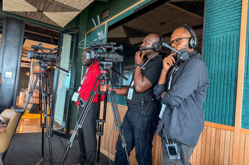 A multi-camera production setup featuring three Mint Glint Media operators manning two professional Sony cameras on tripods. They are all wearing headsets, communicating, and looking attentively toward the filming area in a modern room with green acoustic wall panels.