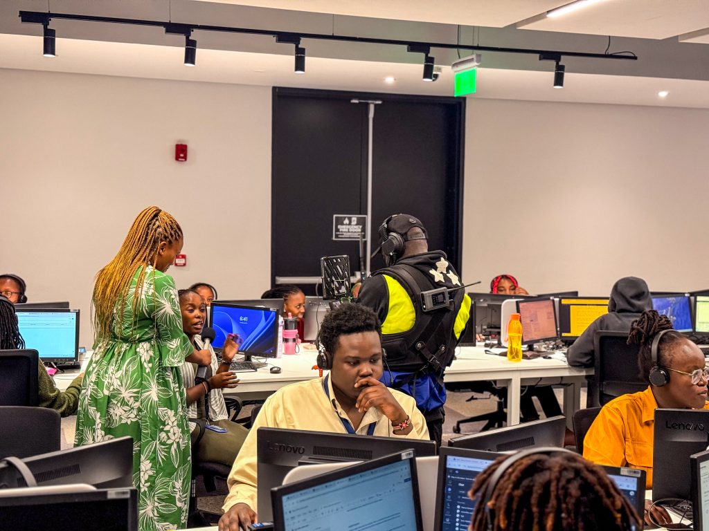A Mint Glint Media camera operator wearing a stabilization rig films a woman in a green dress conducting an interview with an employee at a computer desk in a busy corporate office.